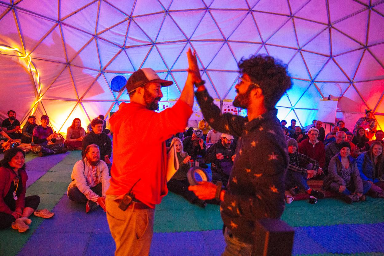 Two people are high fiving in a colorful dome, as an audience looks on. This is a talent show where one man is the emcee and the other younger man is a performer.