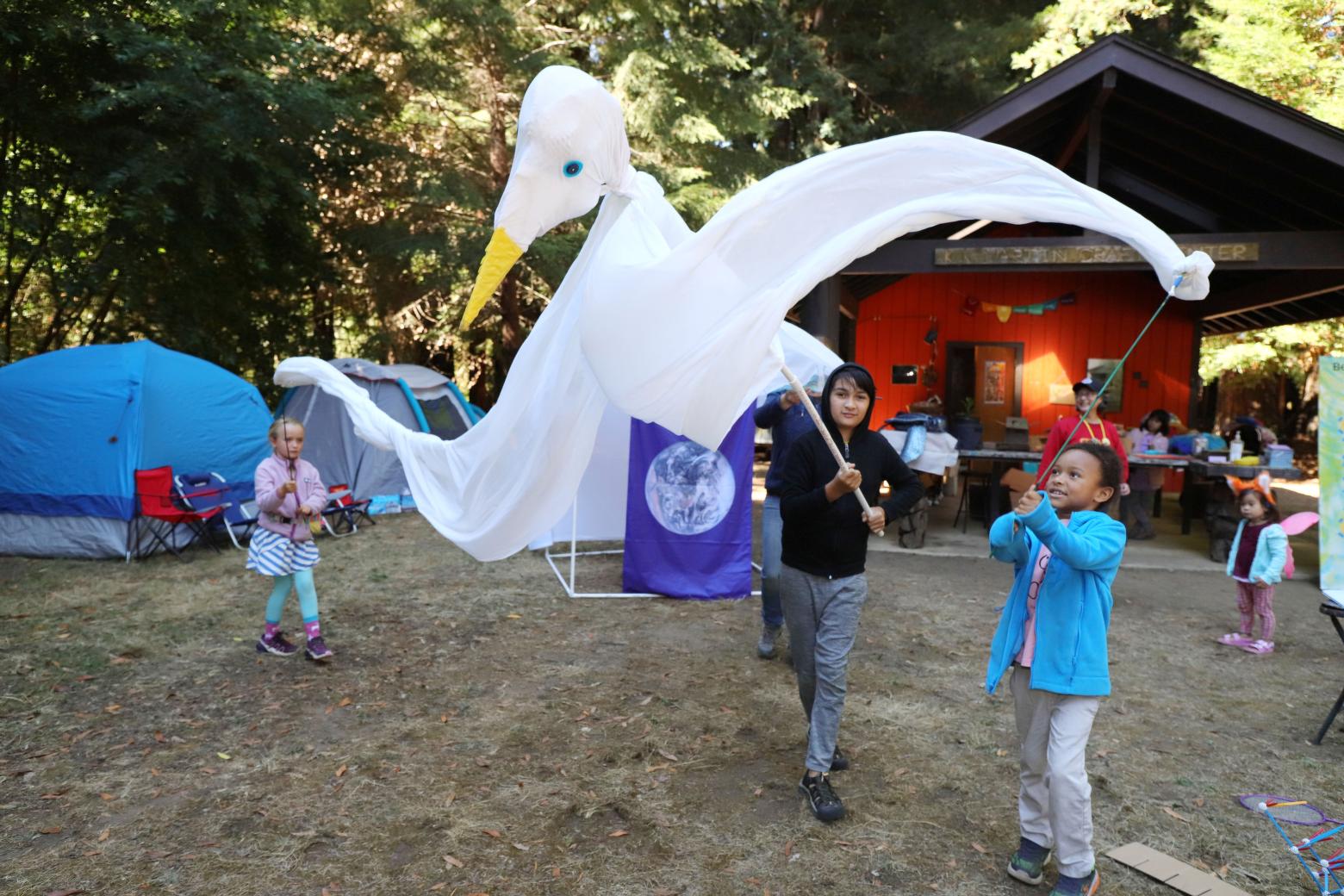 Children holding sticks hold up large bird puppet as they parade through camp grounds.